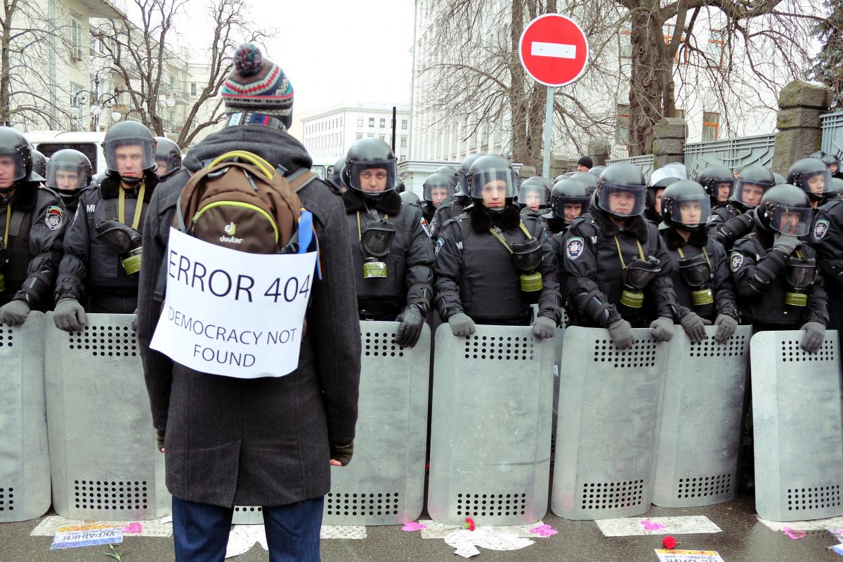 1 грудня 2013 року. Міліція охороняє прохід до Адміністрації Президента. На передньому плані – юрист Сергій Бабенко, учасник Революції Гідності з Полтавщини. На плакаті написано: "Помилка 404. Демократію не знайдено"